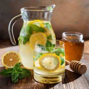 A glass pitcher and glasses filled with fresh mint lemonade, garnished with lemon slices, mint leaves, and honey on a rustic wooden table.